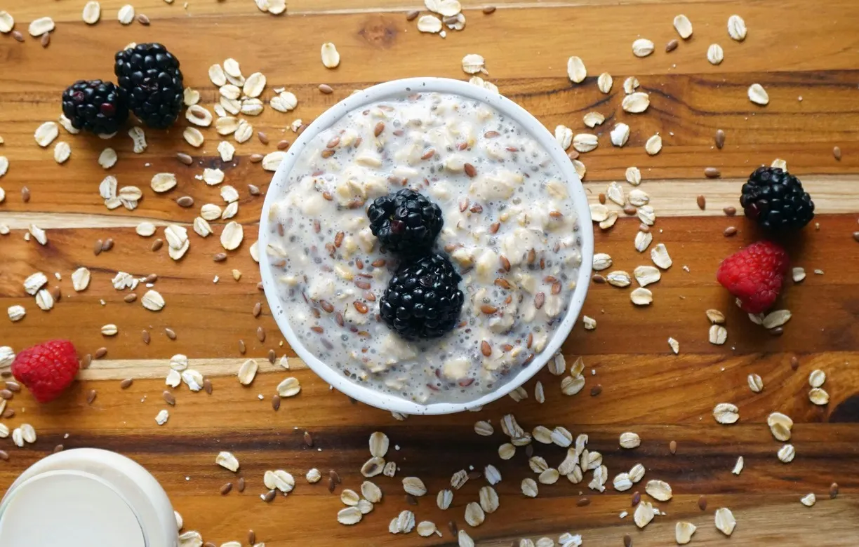 Bowl of overnight oats topped with blackberries on a wooden table with scattered oats and raspberries